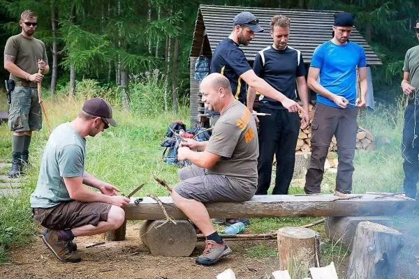 Zwei Personen bearbeiten Holz auf einer Bank, während andere mit Seilen arbeiten