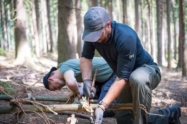 Zwei Personen bearbeiten Holzstücke im Wald, einer schnitzt mit einem Messer