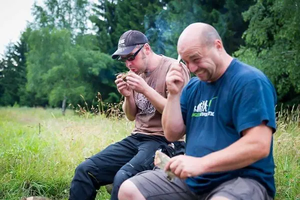Zwei Personen sitzen im Freien und halten rauchende Holzstücke in der Hand