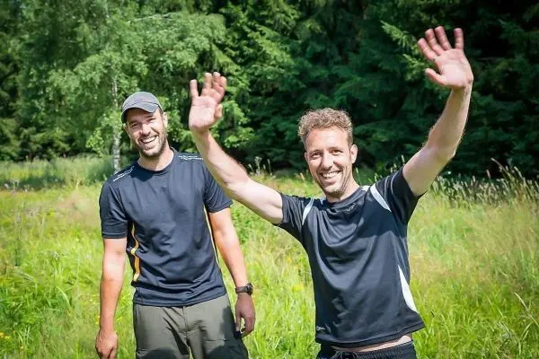 Zwei Personen stehen in einer Wiese und winken freundlich