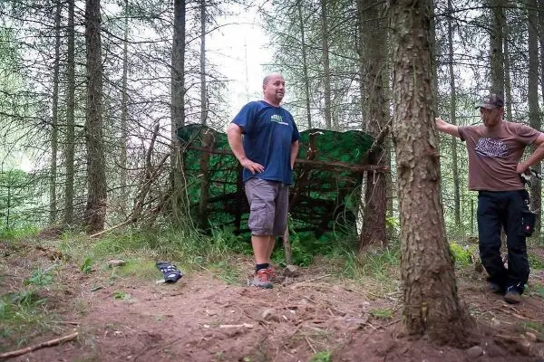 Zwei Personen stehen neben einer selbstgebauten Tarp-Notunterkunft im Wald