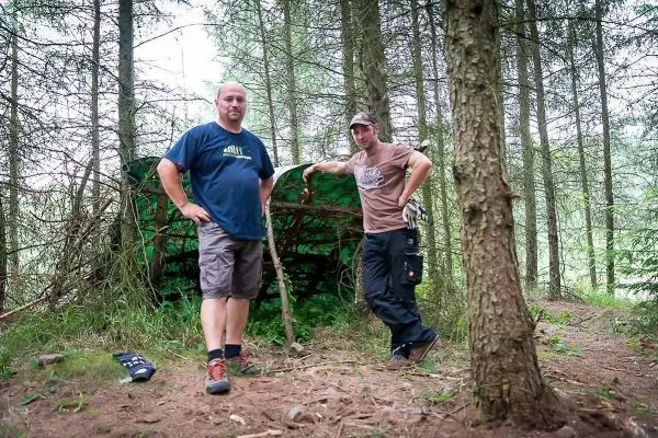 Zwei Personen stehen neben einer selbstgebauten Tarp-Notunterkunft im Wald