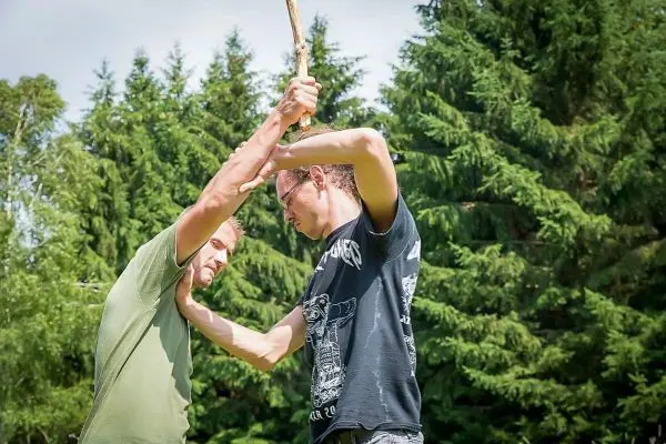 Zwei Personen üben eine Technik mit einem Stock im Freien unter Bäumen