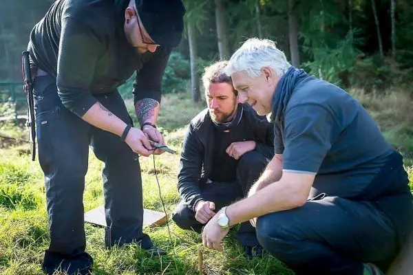 Drei Personen betrachten ein Smartphone und arbeiten an einem Holzstock im Gras