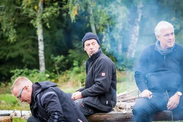 Drei Personen sitzen auf einem Baumstamm in der Natur, Rauch steigt auf