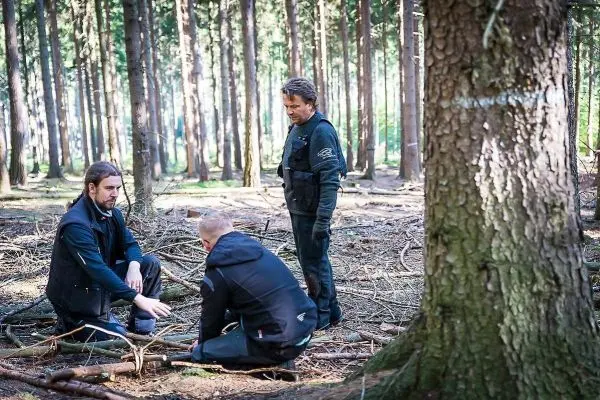 Drei Personen sitzen und stehen im Wald, einer untersucht den Boden