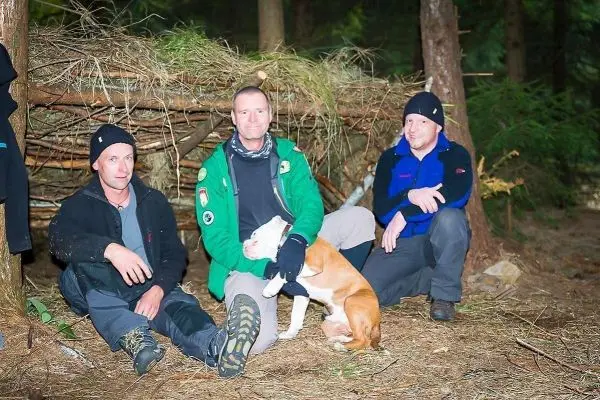 Drei Personen sitzen vor einer selbstgebauten Unterkunft aus Ästen im Wald