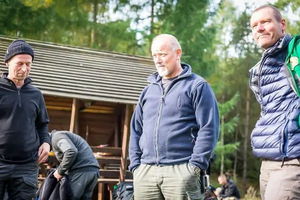 Drei Personen stehen vor einer Holzhütte im Wald, eine Person trägt eine Jacke