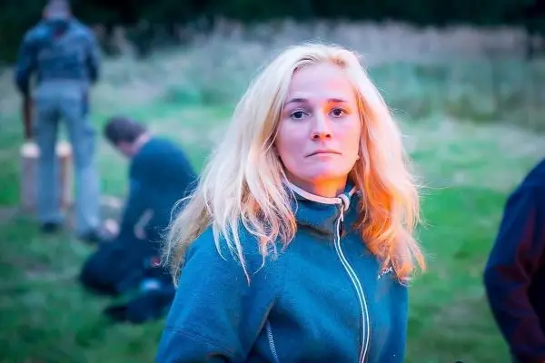 Frau mit blonden Haaren in blauer Jacke steht im Vordergrund, andere Personen im Hintergrund