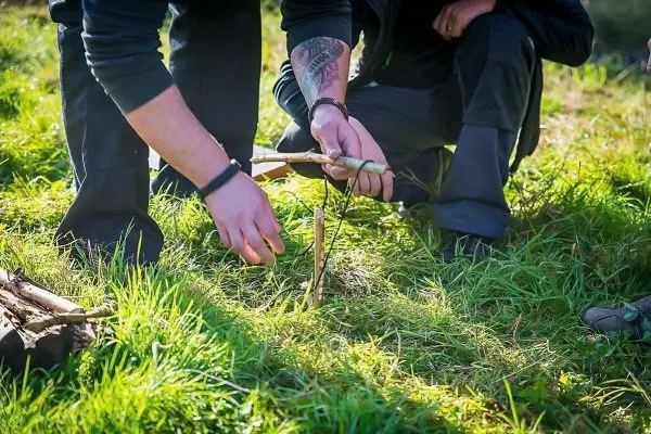 Hände formen eine Schlagfalle (Figure-Four) aus Holzstücken im Gras