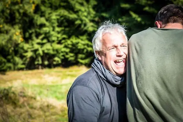 Lachender Mann steht neben einer Person im Freien, umgeben von Bäumen