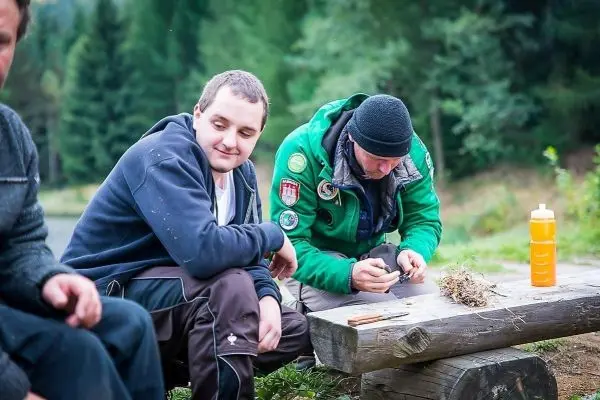 Mann bearbeitet Zunder auf einem Holzstamm, während ein anderer zuschaut