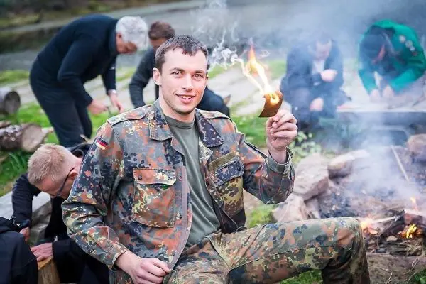 Mann hält brennendes Zundernest über Feuerstelle, im Hintergrund weitere Personen