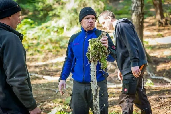 Mann hält einen mit Moos bewachsenen Stock in der Hand, andere Personen im Hintergrund