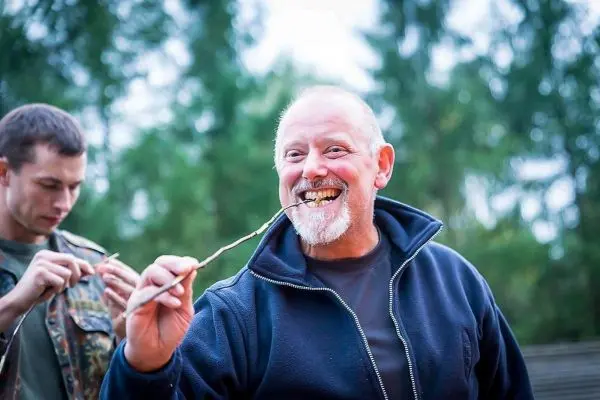 Mann hält einen Stock im Mund und lächelt, während ein anderer Mann im Hintergrund arbeitet