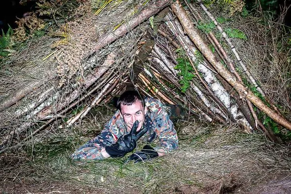 Mann in Tarnkleidung liegt in einer selbstgebauten Laubhütte aus Ästen und Gras