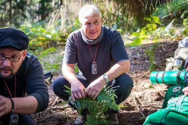 Mann kniet im Wald und betrachtet Farne, Rucksack und Ausrüstung im Hintergrund