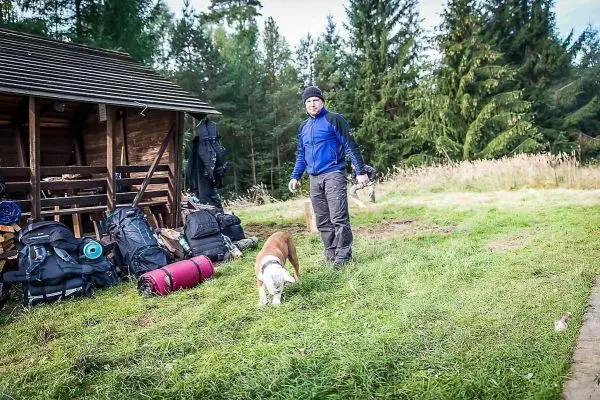 Mann mit Hund steht neben Rucksack und Holzunterstand im Wald