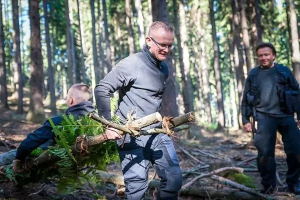 Mann trägt einen Ast mit Farnen durch den Wald, zwei weitere Personen im Hintergrund