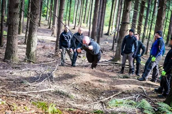 Mann überquert selbstgebaute Seilbrücke zwischen Bäumen, Zuschauer beobachten