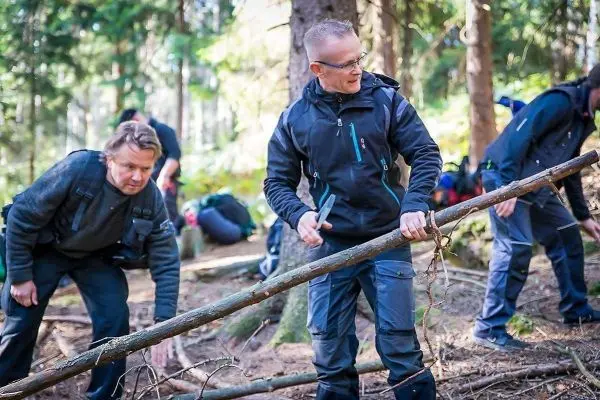 Messer bearbeitet einen Holzstock im Wald, während andere Holz sammeln