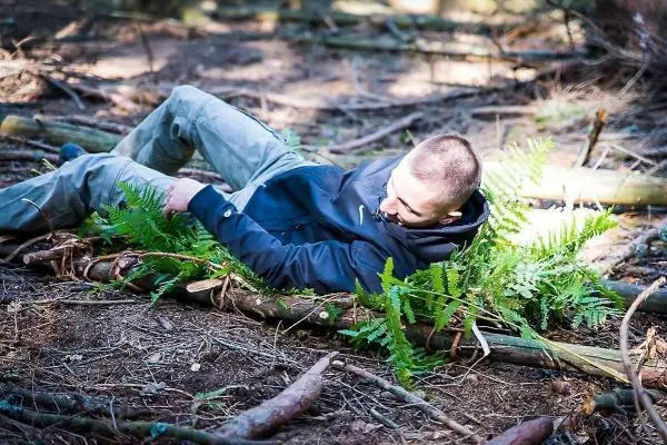 Person liegt auf einem Untergrund aus Ästen und Farnen