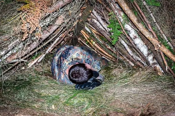 Person liegt in einer selbstgebauten Tarp-Notunterkunft aus Ästen und Gras