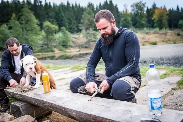 Person schnitzt mit einem Messer an einem Holzstück, Hund sitzt daneben