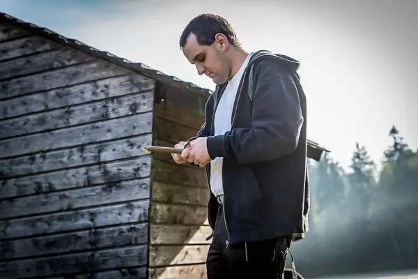 Person steht vor einer Holzhütte und schaut auf ein Blatt Papier