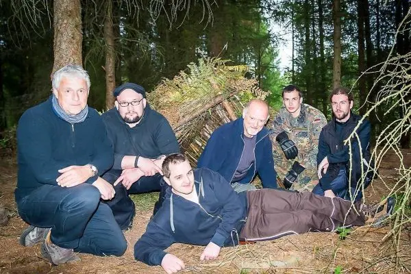 Sechs Personen posieren vor einer selbstgebauten Unterkunft im Wald