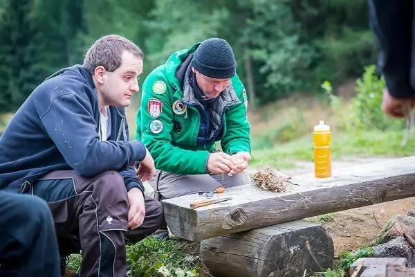 Zunder wird auf einem Holzstück vorbereitet, während zwei Personen aufmerksam zuschauen