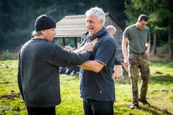Zwei Männer stehen sich gegenüber und interagieren, im Hintergrund weitere Personen und eine Holzstruktur