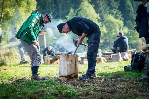 Zwei Personen bearbeiten Holz mit einer Axt auf einer Wiese