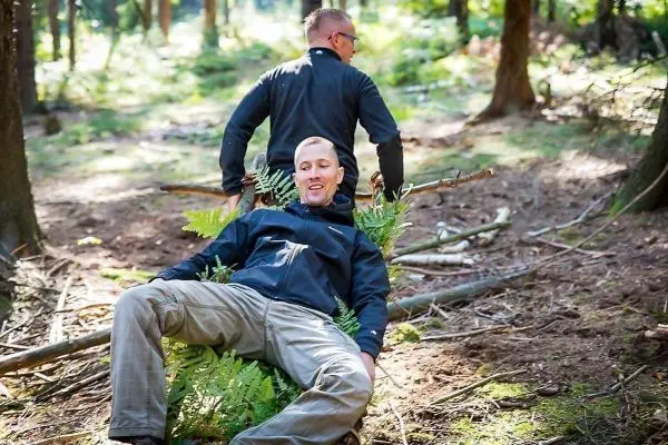 Zwei Personen nutzen eine improvisierte Trage aus Ästen und Farnen im Wald