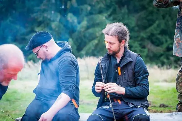 Zwei Personen sitzen auf einer Holzbank und bearbeiten Äste mit einem Messer