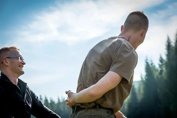 Zwei Personen stehen im Freien, eine Person lacht, während die andere mit hinter dem Rücken verschränkten Händen steht