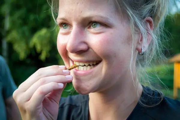 Frau hält einen essbaren Insektensnack zwischen den Fingern und lächelt
