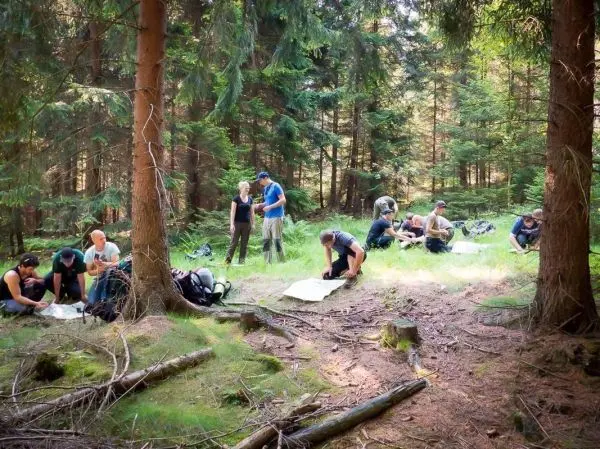 Gruppe arbeitet mit Karten und Rucksäcken im Wald auf moosbedecktem Boden