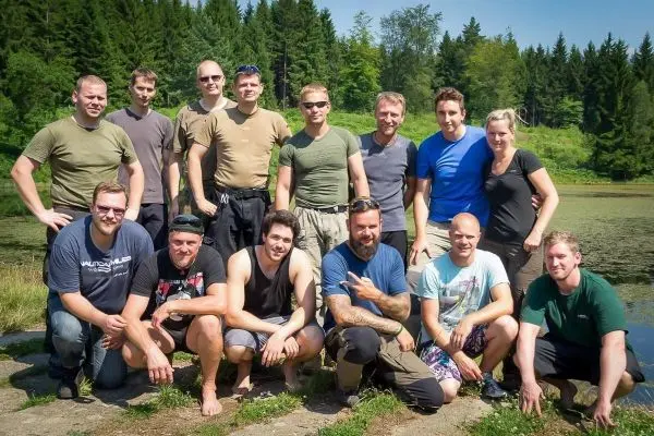 Gruppierung von Personen am Ufer eines Gewässers, umgeben von Bäumen