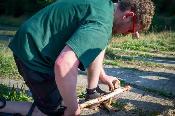 Mann bearbeitet Holzstück mit einem Feuerbohrer zur Feuererzeugung