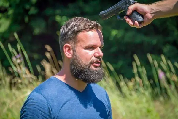 Mann mit Bart steht im Freien, während eine Hand mit einer Pistole auf ihn zeigt