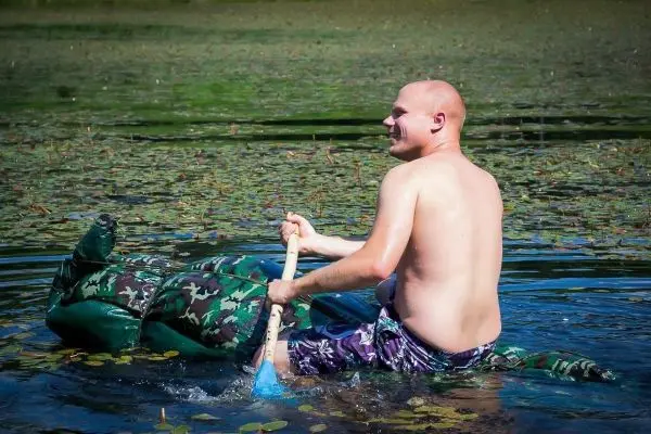 Mann paddelt mit einem Holzpaddel in einem improvisierten Boot aus grünen Materialien