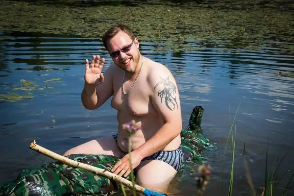 Mann sitzt auf einem camouflagefarbenen Objekt im Wasser und winkt
