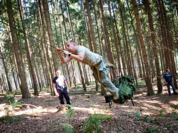 Mann überquert selbstgebaute Seilbrücke im Wald, Rucksack hängt an der Seite
