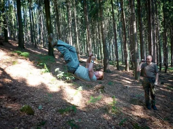 Mann überquert selbstgebaute Seilbrücke im Wald, während ein anderer zuschaut