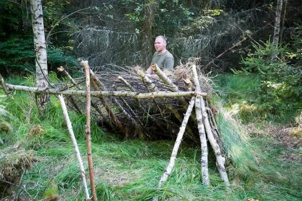Person steht neben einer selbstgebauten Unterkunft aus Ästen und Zweigen