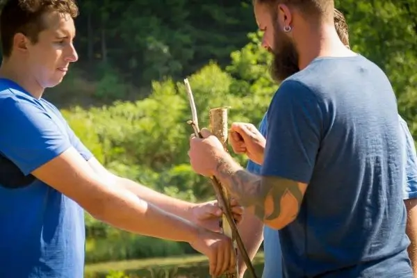 Zwei Personen halten einen Stock, während eine dritte Person einen weiteren Stock hinzufügt