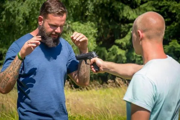 Zwei Personen in einem Freiluft-Szenario, eine Person hält eine Pistole auf die andere