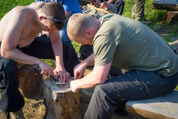 Zwei Personen schnitzen mit Messern auf einem Holzstamm