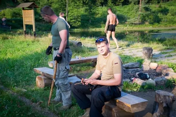 Zwei Personen sitzen auf einer Holzbank, eine bearbeitet ein Stück Holz
